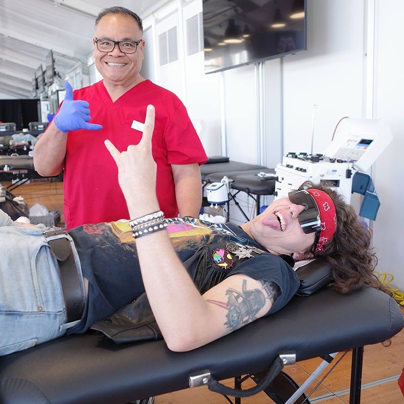 Red Cross nurse standing next to a blood donor, who is lying on a bench in a blood donation room, give a rock and roll hand gesture to the camera.
