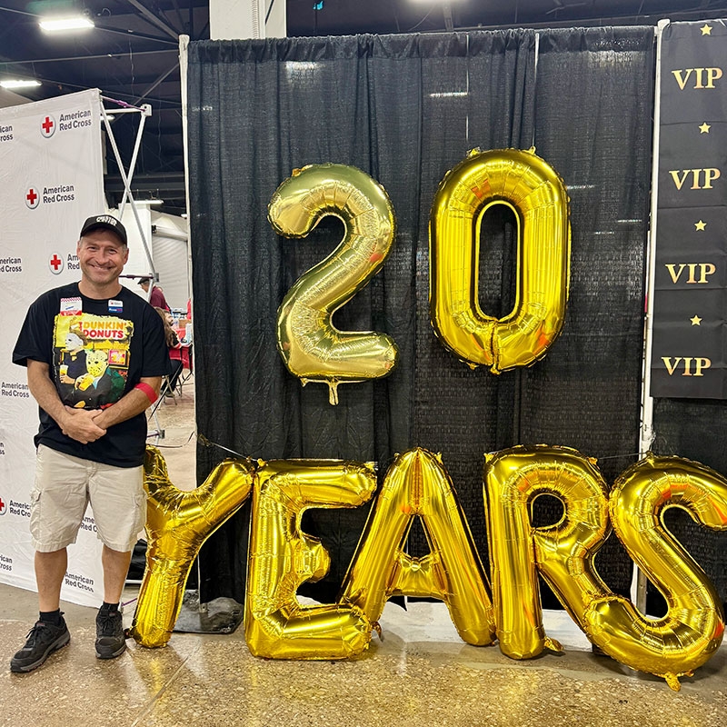 blood donor Tom VanDuyne stands next to balloons that spell out 20 years.
