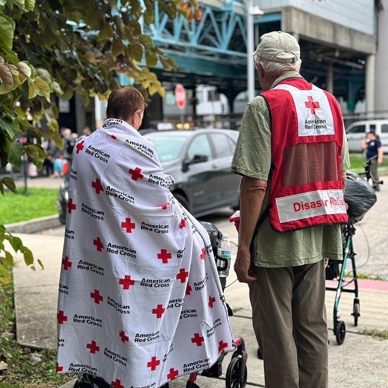 Red Cross volunteer standing outside next to person wearing a Red Cross blanket.