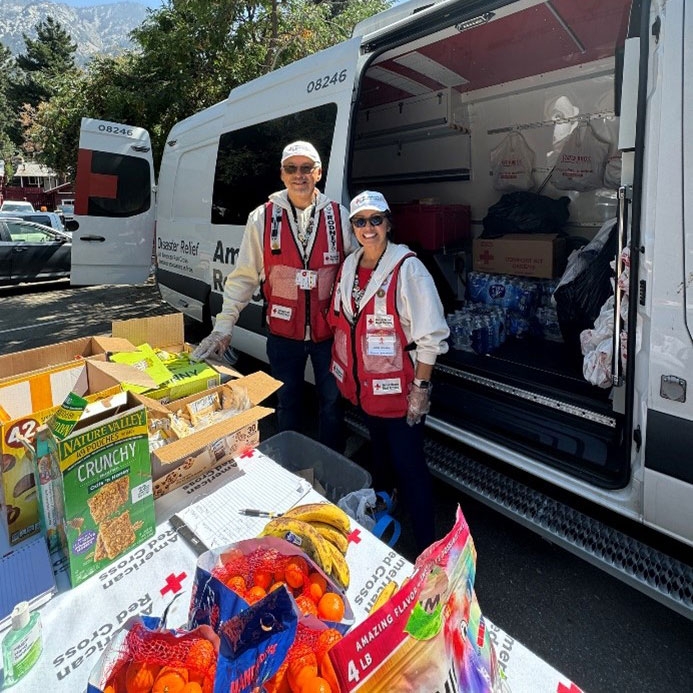 Two Red Cross volunteers standing next to a Red Cross van and a table with packaged food.