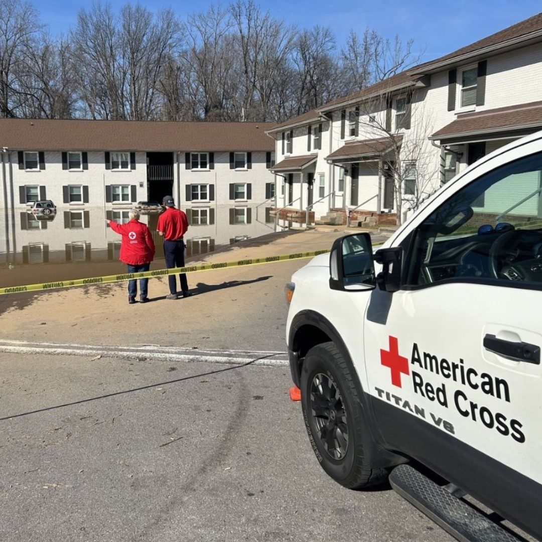 Two red cross volunteers pointing at a flooded area with a red cross vehicle in the background.