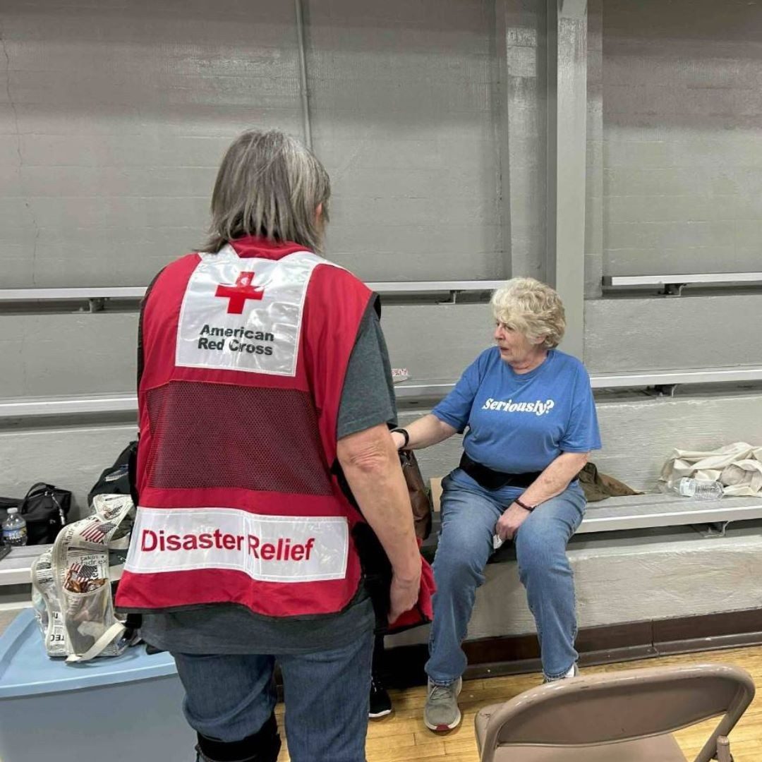 Red cross volunteer wearing a red cross volunteer vest talking to a woman in shelter.