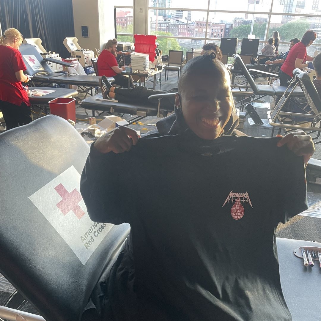 Jamilah holding up Metallica shirt smiling after donating blood.