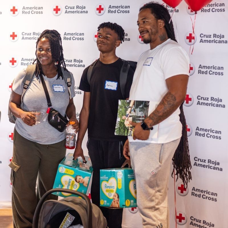 Three people posing for a piture in front of a American Red Cross banner holding diapers in their hand.