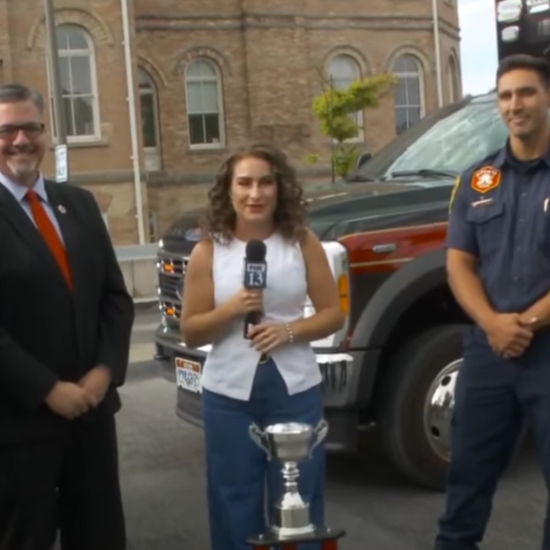 News anchor next to two people with a trophy in the middle. 