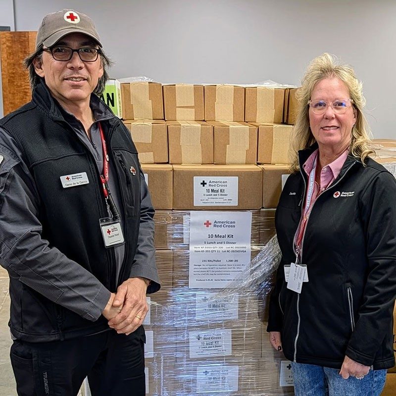 two Red Cross volunteers stand in front of a stack of boxed supplies.