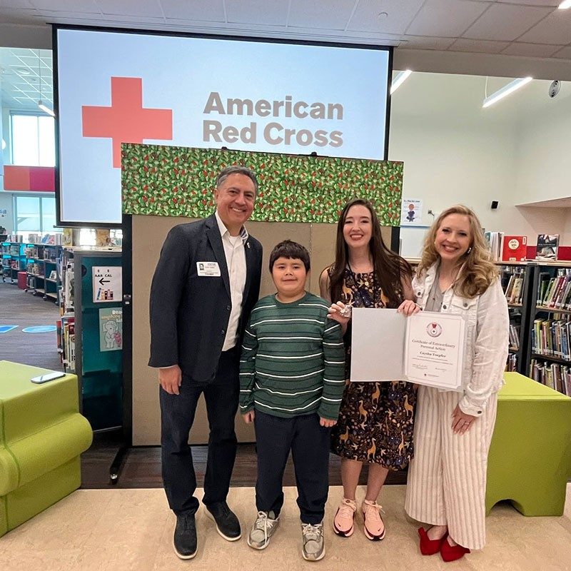 A Red Cross staff member, a young student and another woman standing next to them, presenting the lifesaving award to an elementary school teacher.