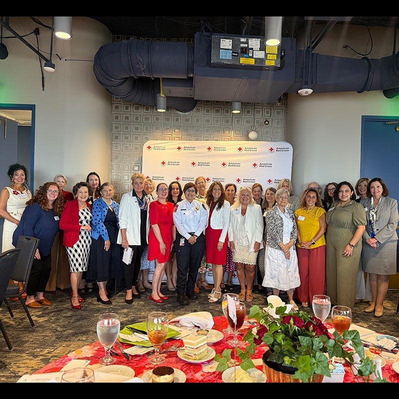 A group of women standing together in front of a Red Cross banner and smiling.