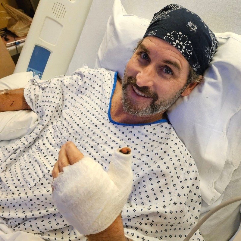 Ben laying in a hospital bed smiling with a bandaged hand.