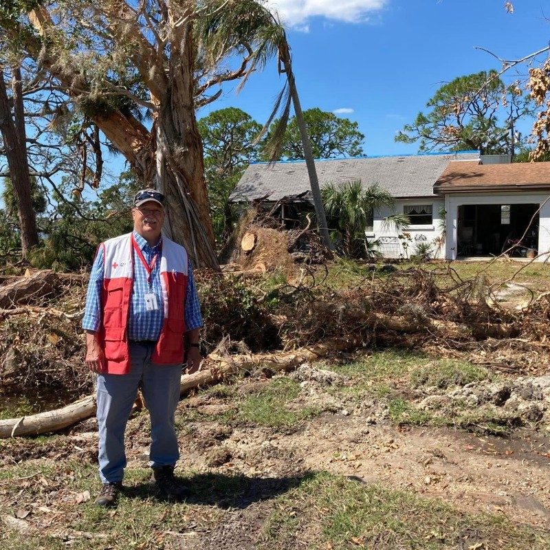 A Red Cross volunteer standing next to a destroyed house.