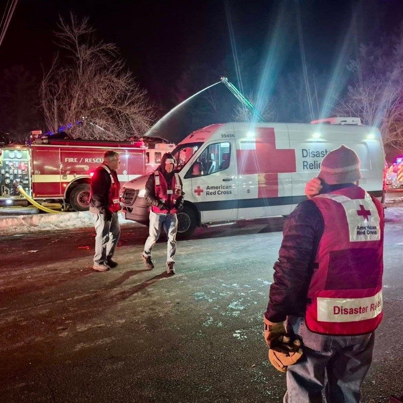 Three Red Cross volunteers in red vest walking towards a fire truck and a Red Cross emergency vehicle.