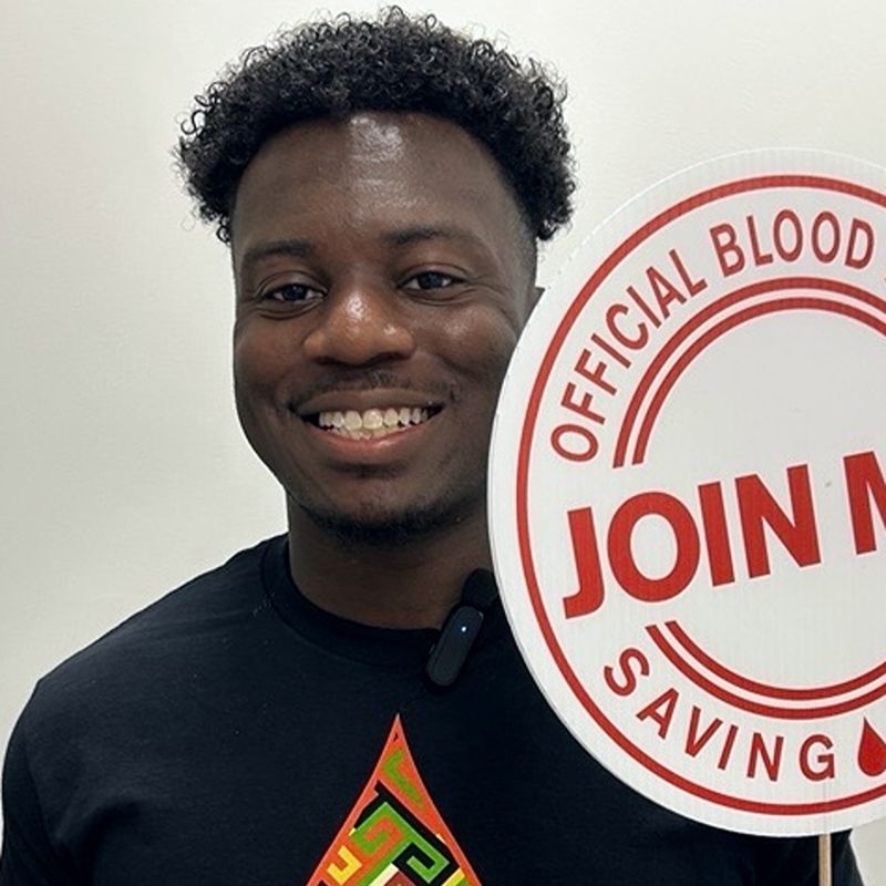 A Red Cross blood donor holding a sign that reads "join me".