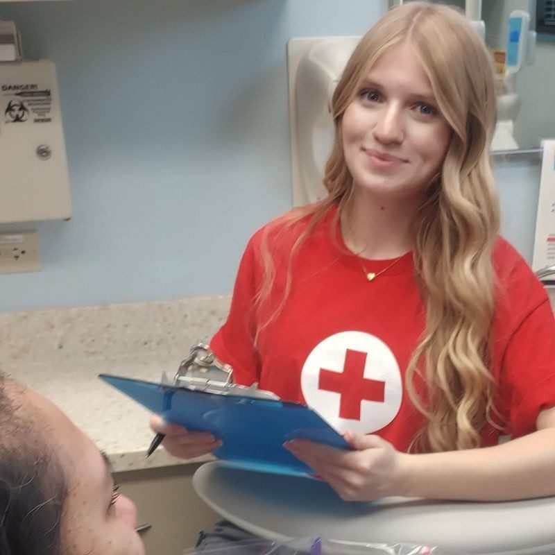 A woman in a Red Cross shirt smiling with a clip board in her hand.