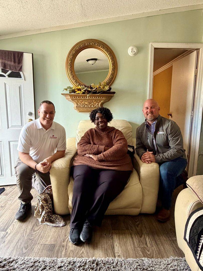 Woman sits on cushioned chair and two men in Red Cross shirts kneel on either side of her