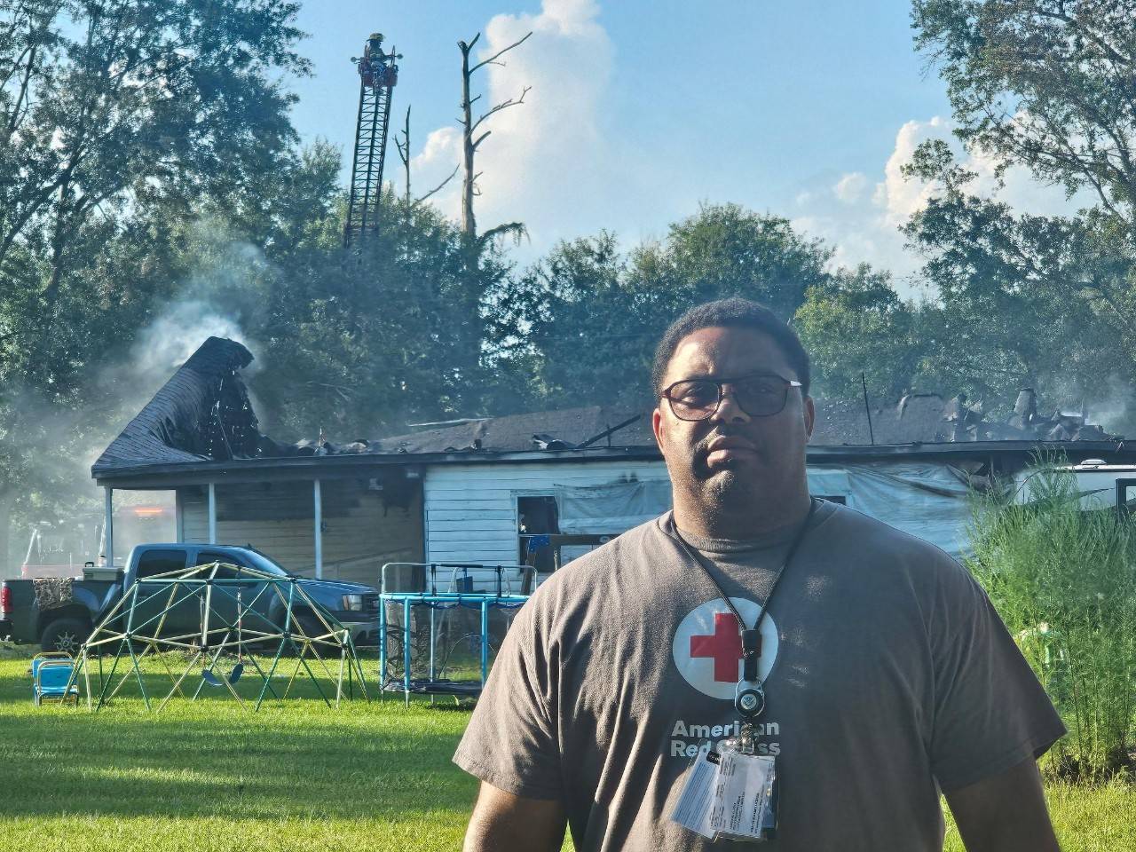 Man in Red Cross shirt stands in front of home with smoke coming from the roof