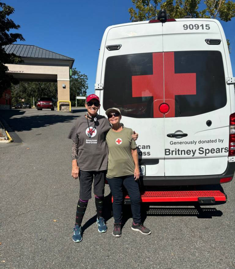 Two women, smiling, stand arm-in-arm in front of an American Red Cross vehicle with a large red cross symbol.