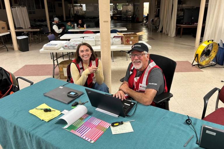 Two Red Cross volunteers sit at table inside REd Cross shelter