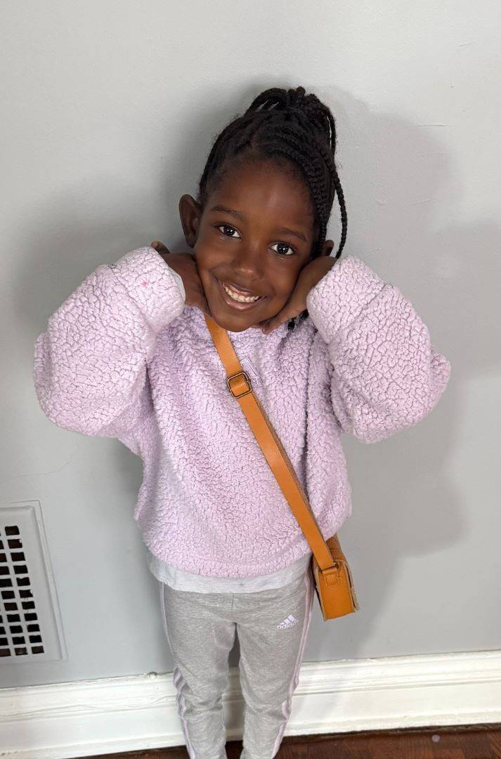 Young girl in pink sweatshirt smiles at camera with hands tucked under her chin