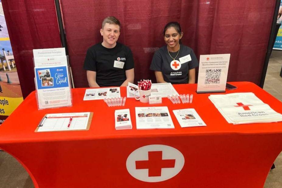 Young woman and man sit behind table with Red Cross tablecloth and resources