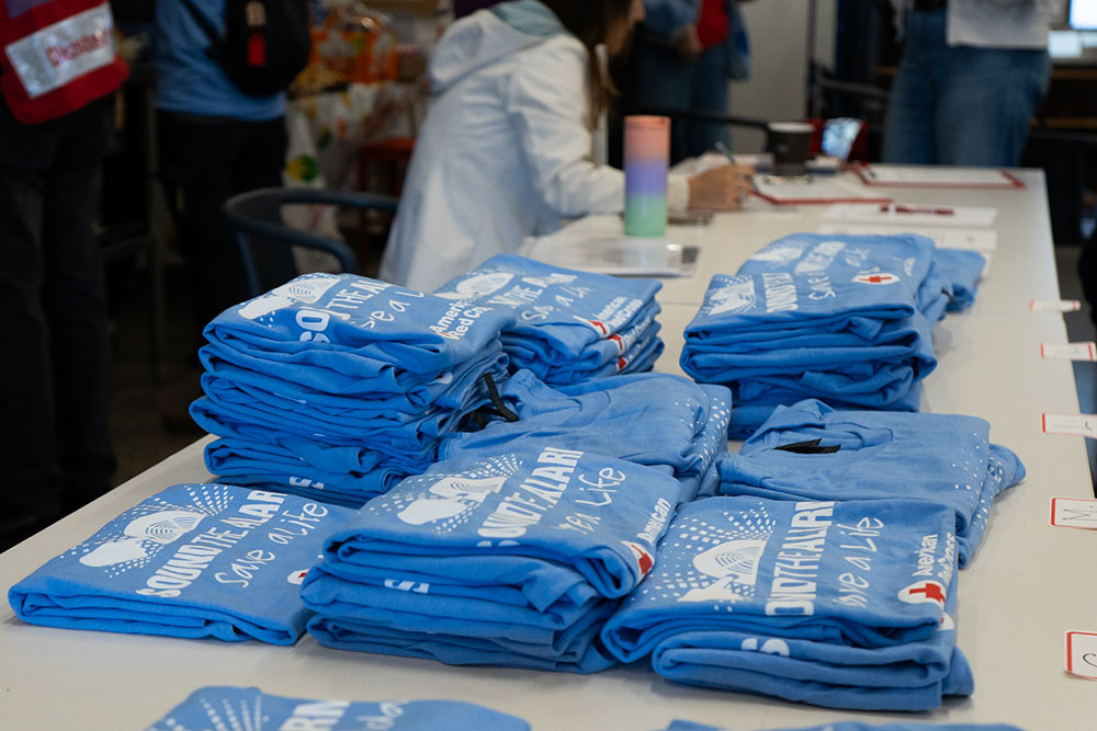 Stacks of folded blue t-shirts with white text, "Sound the Alarm, Save a Life," are on a table at an event. People are in the background.