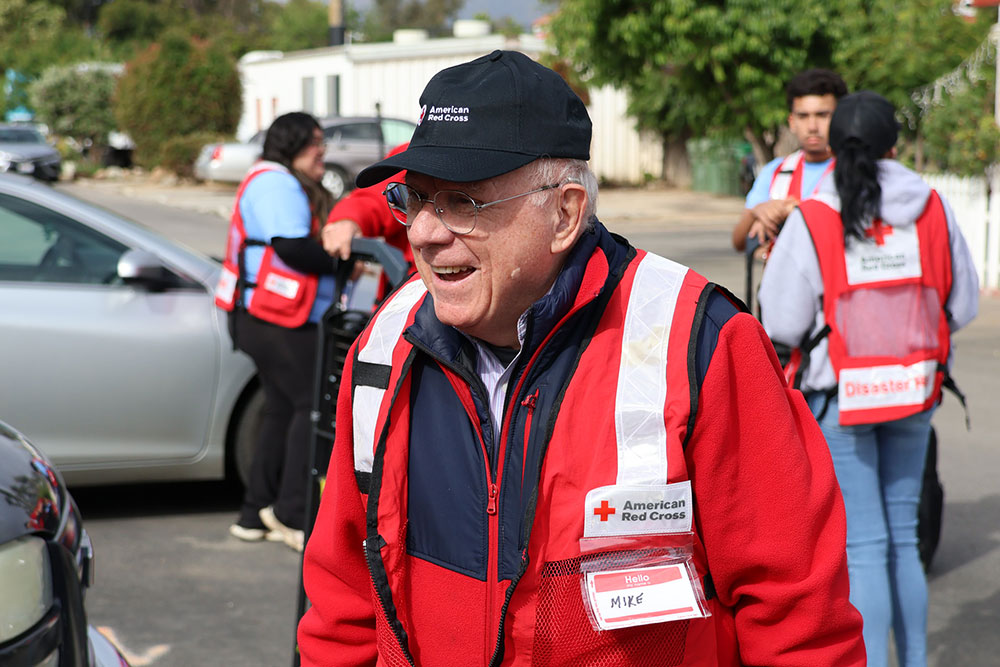 Smiling elderly man in a Red Cross vest and cap stands outdoors, surrounded by volunteers in similar attire.