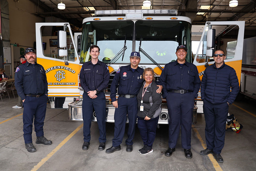 A group of six people, some in uniforms, stand smiling in front of a yellow fire truck in a station.