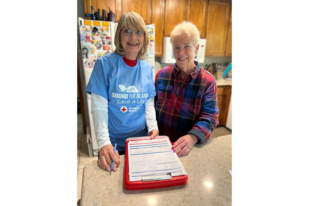 Two smiling women stand in a kitchen. One wears a "Sound the Alarm" shirt and holds a clipboard.