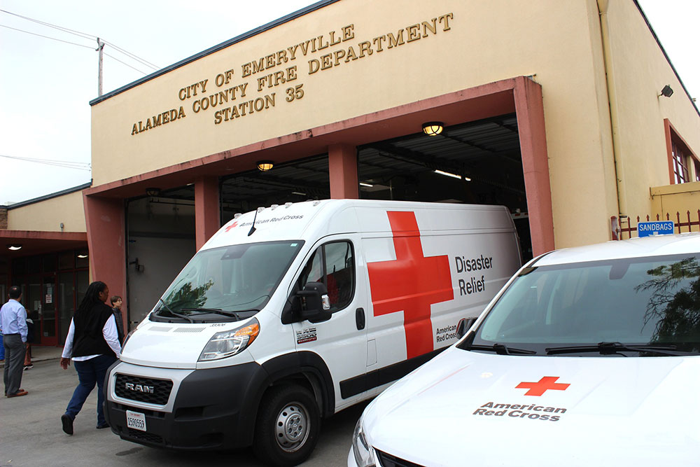 Two American Red Cross vans, marked for disaster relief, are parked outside the City of Emeryville Alameda County Fire Department Station 35. People walk nearby.