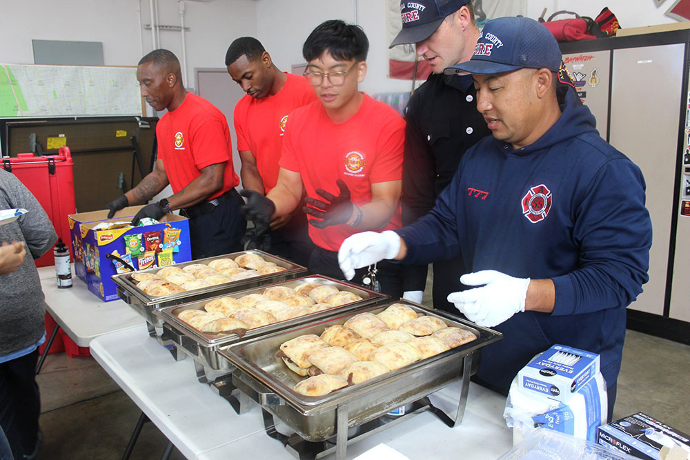 Firefighters in red and blue uniforms serve food from trays.
