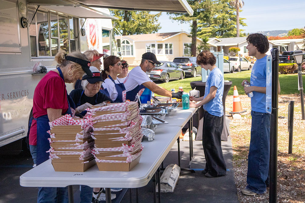 Volunteers serve meals from a mobile food truck, with stacks of packed lunches on a table. The scene is outdoors on a sunny day.