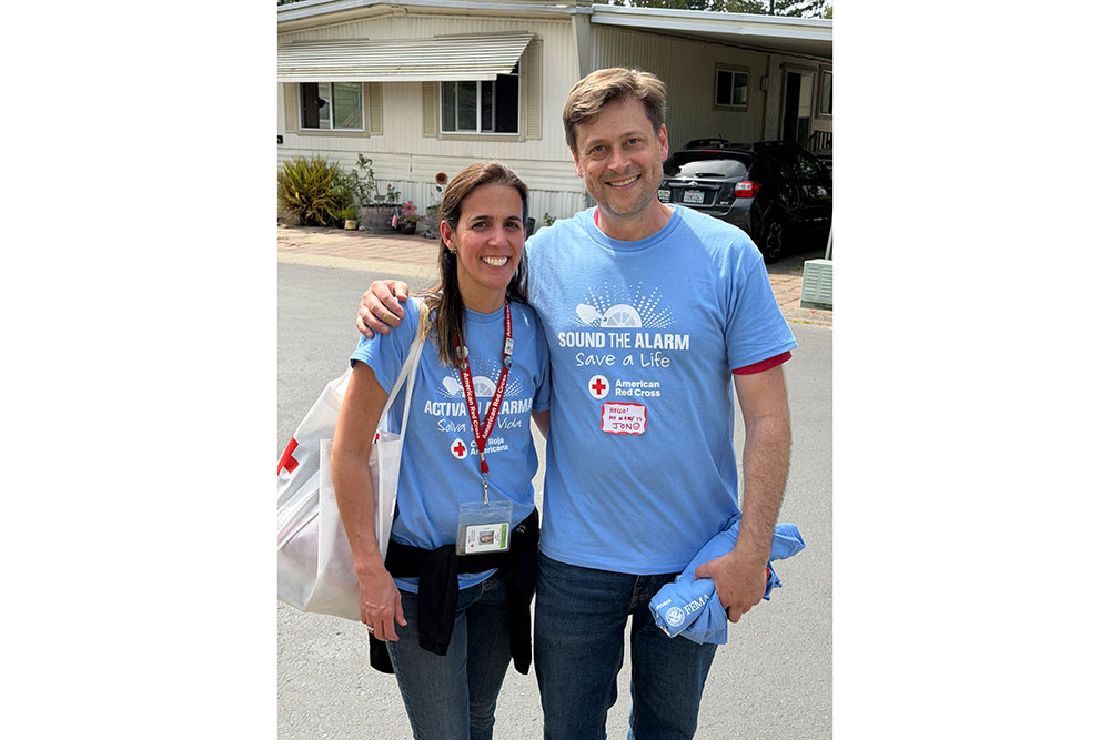 A smiling man and woman in matching blue "Sound the Alarm" Red Cross shirts stand arm-in-arm on a residential street.