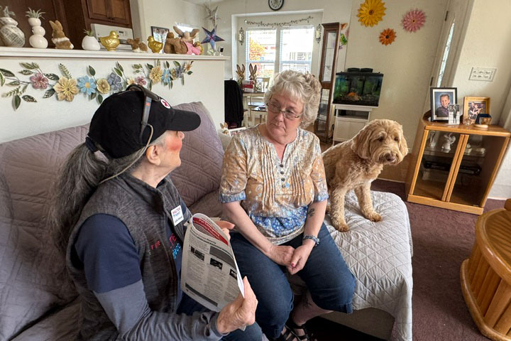 Two women sit on a couch in a cozy living room; one holds a brochure. A dog sits beside them. The room has floral decor and framed photos.