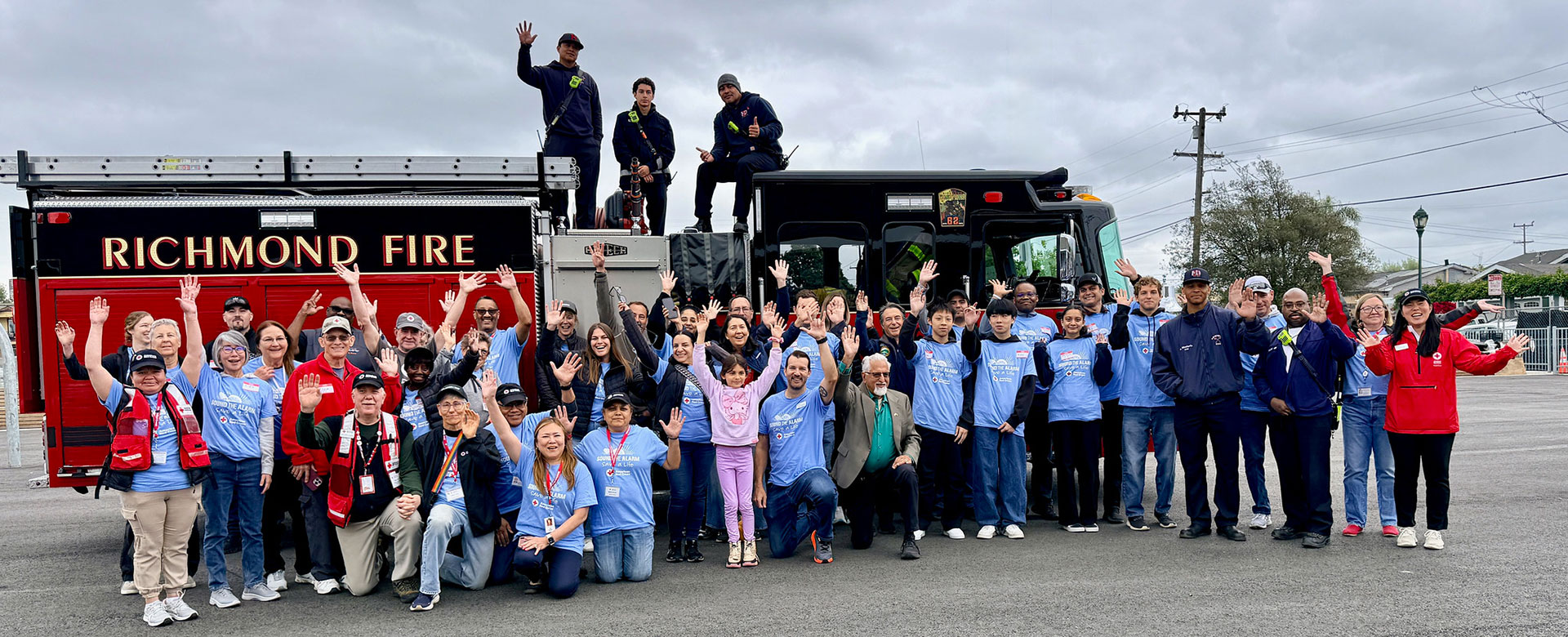 A diverse group of people in casual clothes cheerfully pose in front of a Richmond Fire truck. Some firefighters are on the truck, and the sky is overcast.