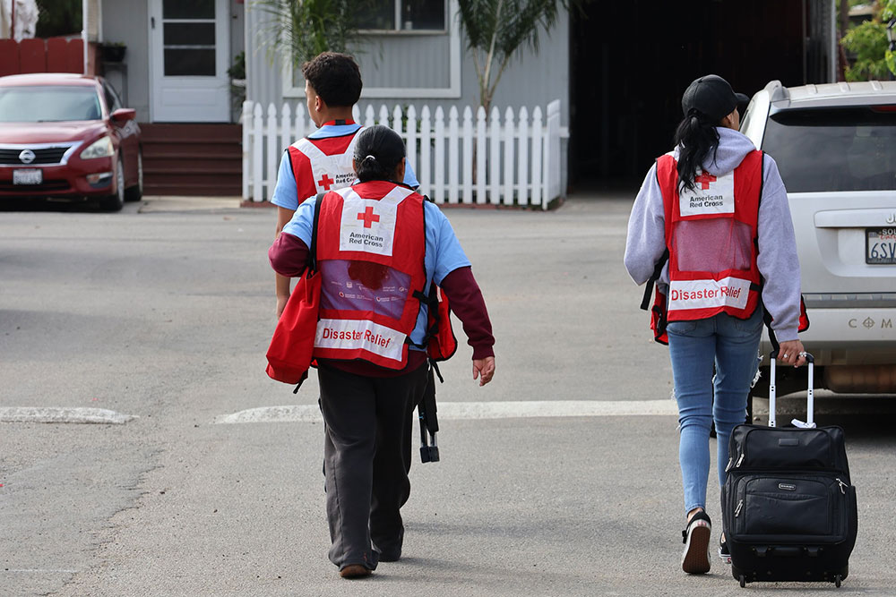 Three Red Cross disaster relief volunteers, wearing red vests, walk away with a suitcase on a residential street.