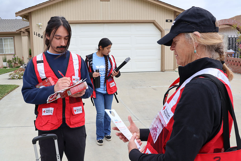 Three Red Cross volunteers prepare for community outreach outside a suburban house. One writes on a clipboard, the other holds smoke alarms.