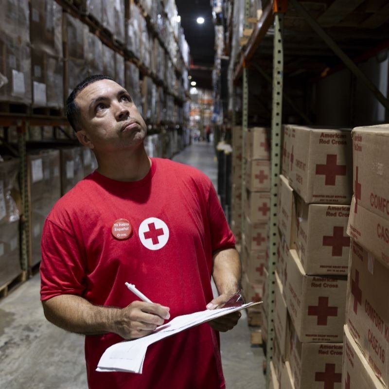 A red Cross volunteer in a warehouse with a clipboard and pens