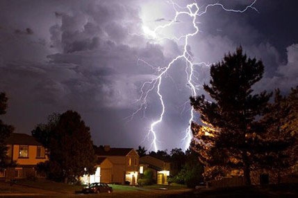 Lightning strikes illuminate a residential area, highlighting homes against a dark stormy sky.