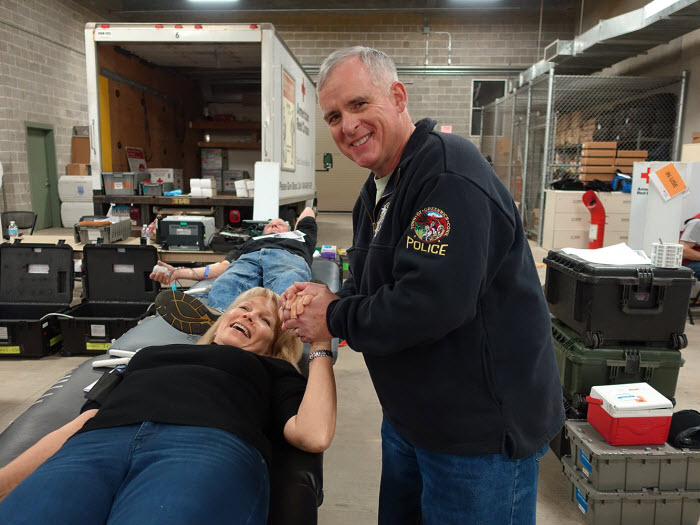 Jim Heavey stands next to a woman lying on a stretcher giving blood and he is holding her hand