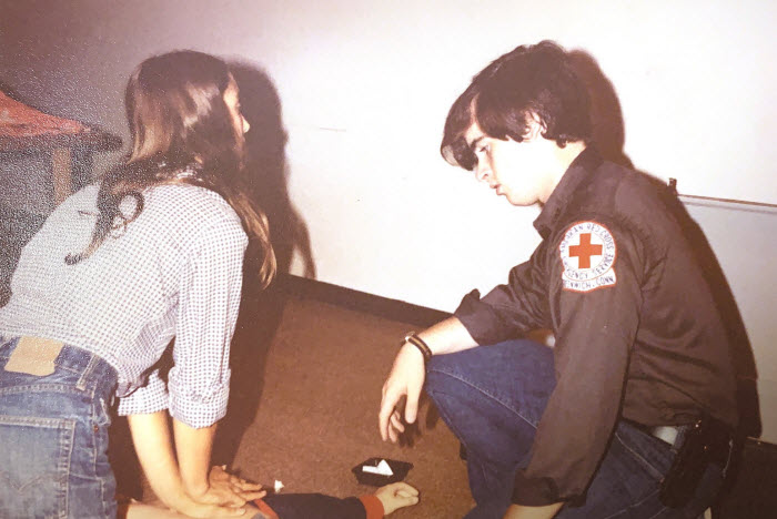 Jim Heavey as a young man kneeling on the ground watching a young woman learn CPR