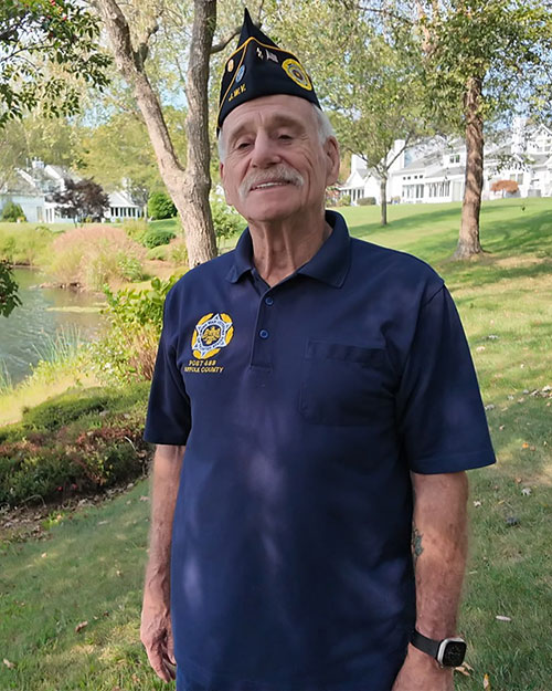 Mel Cohen wearing military hat standing on the grass by a lake.