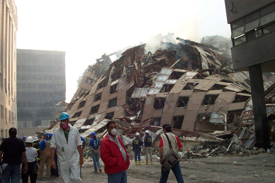 A man in a red jacket and a face mask stands in a dusty street as people walk all around him, with a large pile of building rubble in the background.
