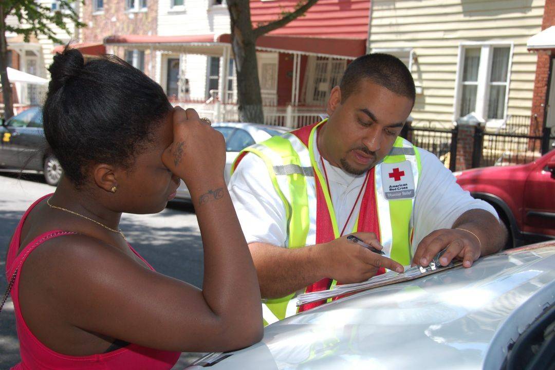 Red Cross disaster worker reviews paperwork with a resident on the hood of a vehicle on a residential street.