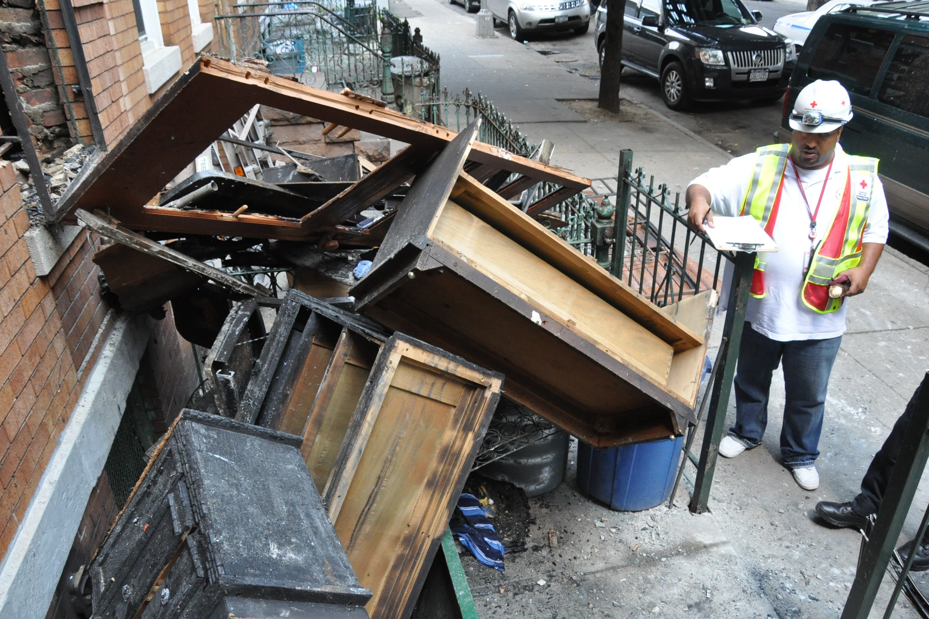 A Red Cross disaster worker surveys fire-damaged furniture and debris piled outside a residential building.