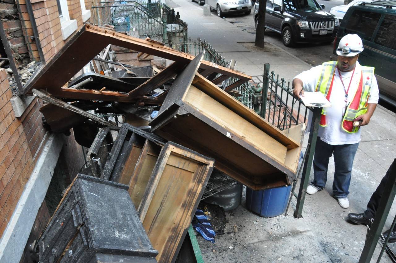 A Red Cross disaster worker surveys fire-damaged furniture and debris piled outside a residential building.