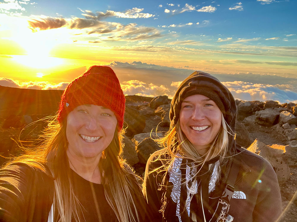 two people wearing beanies and sweaters with the sunrise and clouds in the background.