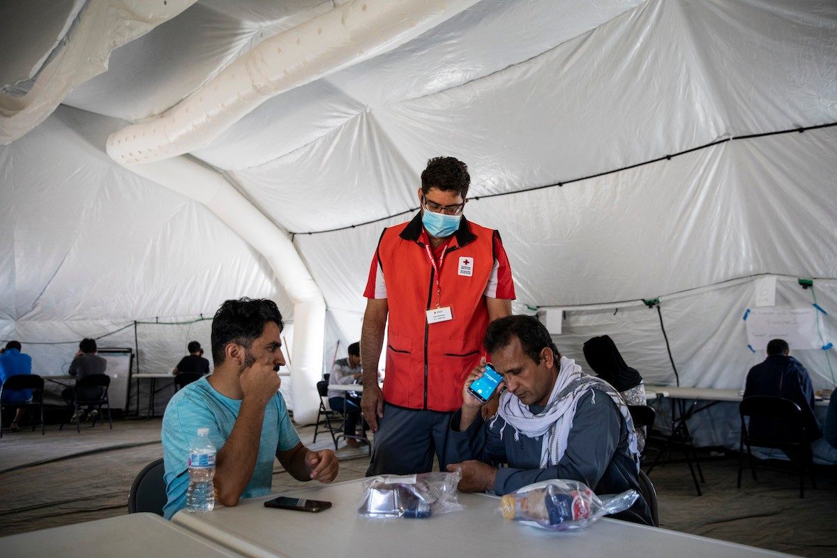 Red cross volunteer wearing mask next to two people sitting at a table.