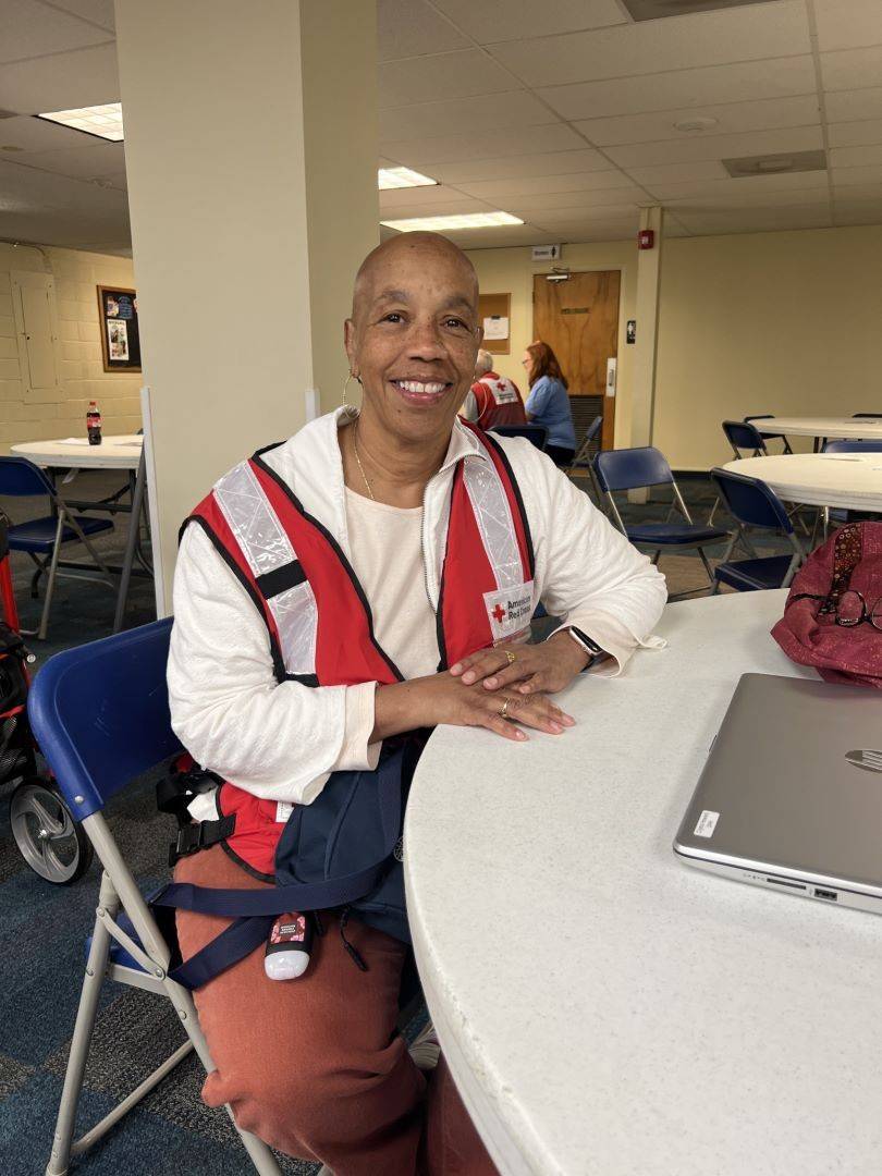 Woman sits at table wearing Red Cross vest and smiling at camera