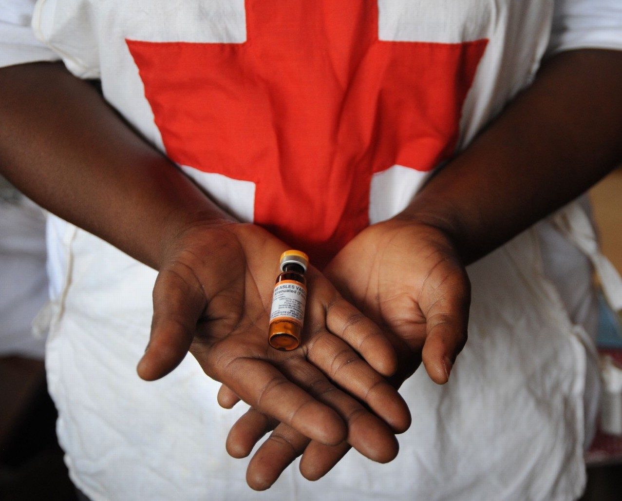 Photo of a person wearing a Red Cross shirt holding a vile of measles vaccine