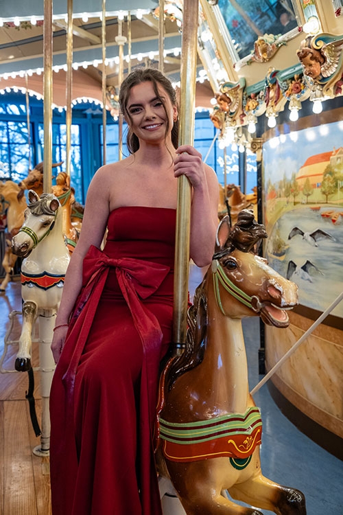 Emily Gindele in a red dress sitting on a merry-go-round and smiling.