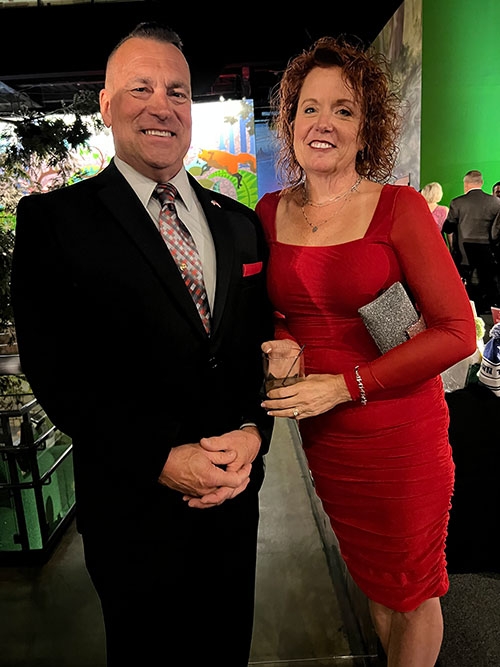 Trooper John Finkbiner wearing a suit and his wife wearing a red dress. both smiling for the picture.